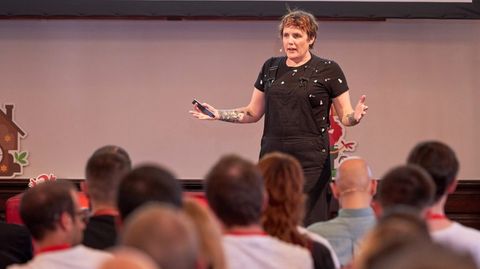 Mia from behind,
standing at a laptop -
speaking to a conference audience
and gesturing to one side
